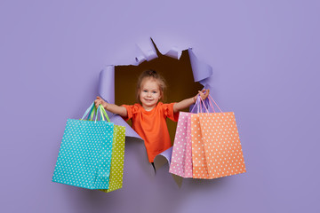 Cute little child girl breaks through a colored purple paper wall with shopping bags in hands. Toddler funny emotions face. Copy space for text