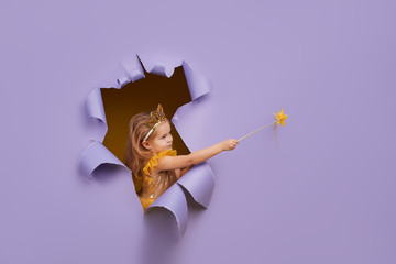 Cute little child girl in princess costume breaks through a colored purple paper wall. Points with a magic wand to the empty space on the right. Toddler funny emotions face. - obrazy, fototapety, plakaty