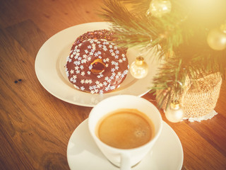 Chocolate donuts sprinkled with colorful stars on the table. Coffee and Christmas decorations in the background. Donut on Christmas table. Chocolate donut at sunrise. Coffee and donut.