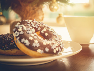 Chocolate donuts sprinkled with colorful stars on the table. Coffee and Christmas decorations in the background. Donut on Christmas table. Chocolate donut at sunrise. Coffee and donut.