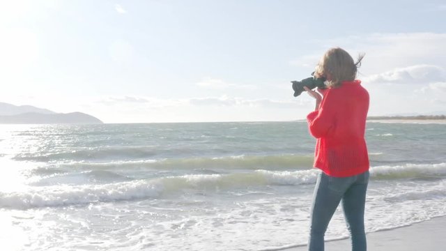Woman with red knitted sweater and DSLR camera taking pictures on the beach, slow motion.
