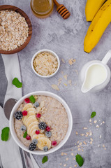Oatmeal in a bowl with banana, frozen berries, almond petals, mint leaves and honey on a gray stone background. Tasty and healthy breakfast. Copy space.