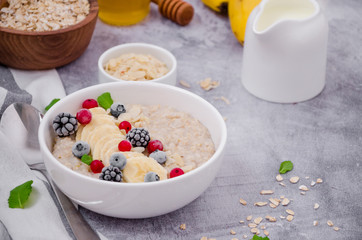 Oatmeal in a bowl with banana, frozen berries, almond petals, mint leaves and honey on a gray stone background. Tasty and healthy breakfast. Copy space.