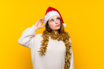 Girl with christmas hat over isolated yellow background having doubts and with confuse face...