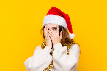 Girl with christmas hat over isolated yellow background covering eyes and looking through fingers
