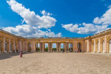 Lovely panoramic view of the cobbled stone courtyard and the sheltered colonnade connecting the two wings of the famous Grand Trianon Palace in Versailles on a nice summer day with a blue sky. - obrazy, fototapety, plakaty