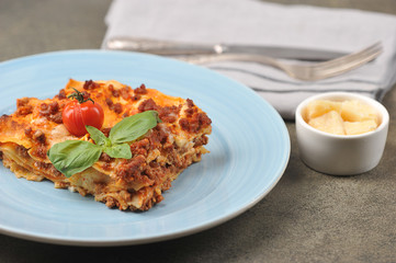 Serving of lasagna on a light plate.  Lasagna garnished with cherry tomatoes and fresh basil leaves.  Next to the plate are cutlery and slices of hard Parmesan cheese.  Close-up.