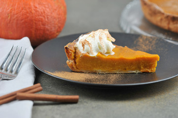 A piece of pumpkin pie on a black plate.  The concept of a festive treat of the autumn harvest.  Close-up.  In the frame, cinnamon sticks, pumpkin, pie.