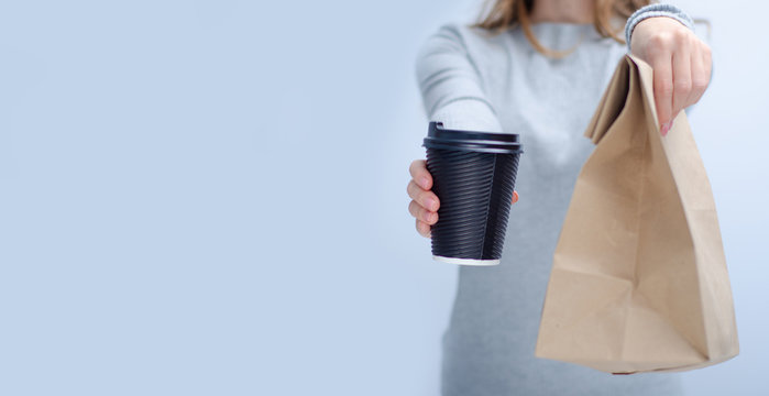 Woman holding cardboard cup of coffee and package food take and go in hand on gray light background