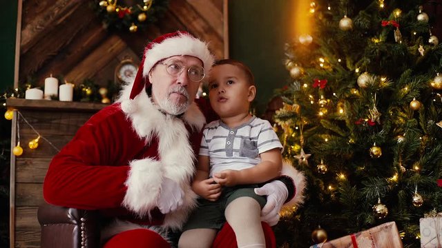 Little boy whispering Santa Claus ear him christmas wish sitting on his laps, next to the Christmas tree in room with festive interior. Shooting in slow motion.