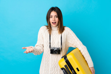 Traveler woman holding a suitcase over isolated blue wall with shocked facial expression