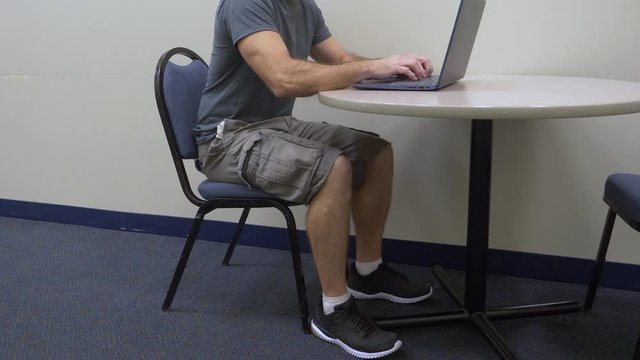 Man sitting on office chair working, typing on a laptop computer at a table in a hallway.