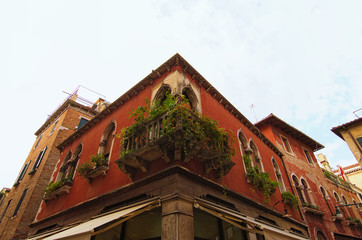 Amazing vintage balcony with many different potted flowers on the corner of medieval red-colored building against blue sky.  Travel and tourism concept. Non-tourist part of Venice, Italy