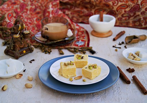 Indian sweets with pistachios, almonds and cardamom on a white plate on a light concrete background. Served with masala tea. Traditional Indian sweets.