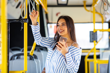 Woman Using Smartphone While Riding In Public Transport. Young adorable joyful woman is standing on the bus using the phone and smiling. Woman Reading Text Message On Bus