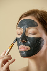 Charcoal face mask or black clay mud. Beautiful woman is applying purifying black mask on her face, close up studio shot. Home spa natural cosmetic concept.