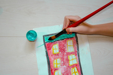 A child draw with paints a red house with windows on a piece of paper.