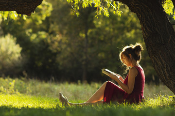 sunny portrait of a beautiful girl sitting on green glade under tree branches with book, woman...