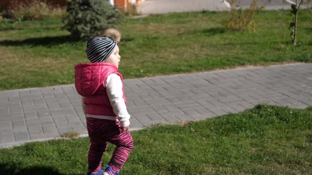  A little girl walks on the grass. A girl walks on the grass. Girl in a pink jacket. Little girl. Stroll. Children. Playground. Sunny day.