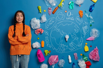 Horizontal shot of pensive Asian woman keeps hands crossed, collects trash and thinks about ecology...