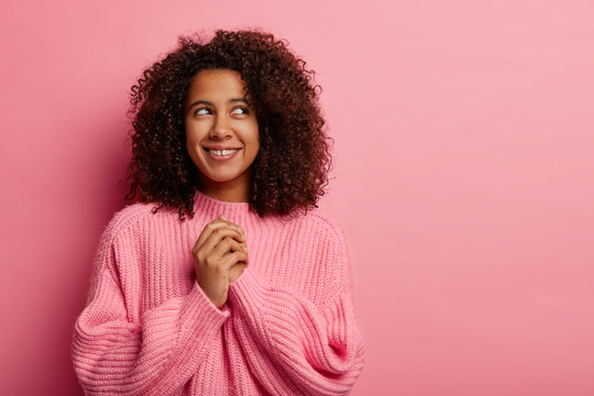 Satisfied dark skinned girl with Afro curly hair keeps hands together, focused above smiles gently wears knitted sweater dreams about something nice has cheerful thoughtful expression isolated on pink
