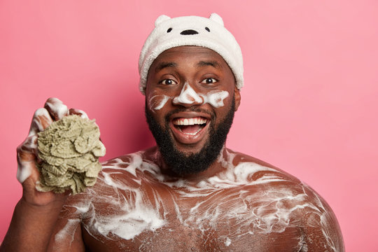 Funny black bearded man washes his torso, has foam on body and face, laughs happily, holds sponge, wears bath hat, isolated on pink background. Naked macho takes shower or douche. Hygiene concept