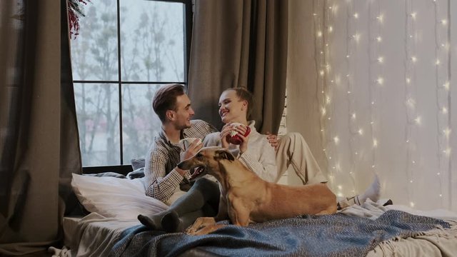 New Year. Young Couple In Cozy Apartment. Couple And Their Dog In Bed. They Drink Tea.