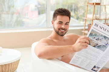 Handsome young man reading newspaper while taking bath at home