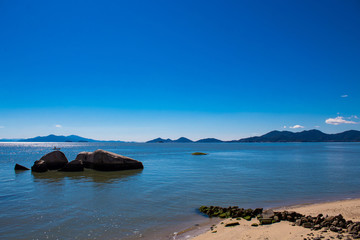  Mar azul tropical em dia de sol, pedras e barco de pesca no fundo montanhas azuis de Florianópolis