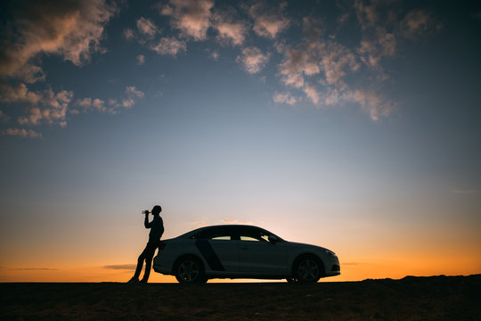 Silhouette Of Man Driver Relaxing After A Ride, Standing Next To His Car And Drinking Water From A Bottle, Side View. Sunset Time. 