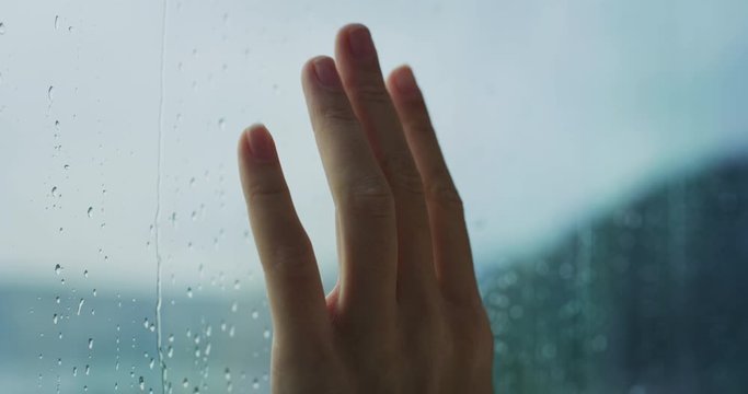 Close up of a young woman hand is touching a glass with water drops while outside is raining.