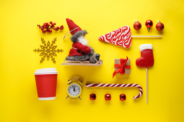 Christmas flat lay composition. Christmas sweets, gift, toys, Santa in a sleigh, an alarm clock with a cool down time and a cup on a yellow background.