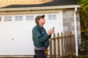 Home inspector looks up at a roof of a property, dictating into software on his smart phone. 