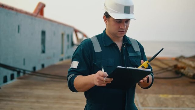 Marine chief officer or chief mate on deck of ship or vessel. He fills up ahts vessel checklist. Ship routine paperwork. He holds VHF walkie-talkie radio in hands.
