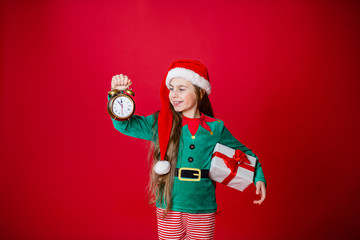 Merry Christmas, happy attractive girl with a clock and gifts in the costume of Santa Claus helper on a bright red bright color background.