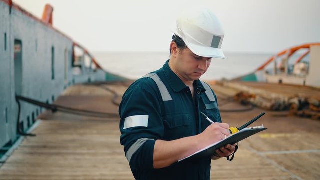 Marine chief officer or chief mate on deck of ship or vessel. He fills up ahts vessel checklist. Ship routine paperwork. He holds VHF walkie-talkie radio in hands.