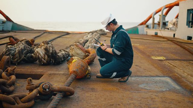 Marine chief officer or chief mate on deck of ship or vessel. He fills up ahts vessel checklist. Ship routine paperwork. He holds VHF walkie-talkie radio in hands.