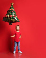 Happy toddler girl in a red T-shirt, blue jeans holding a Christmas tree balloon on a red background with free copy space.