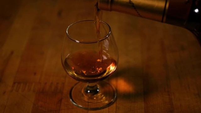 Single snifter glass on wooden table. Cognac out of bottle being poured in.