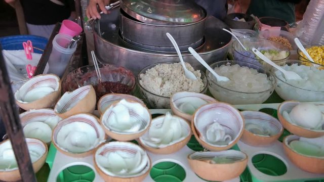 Vendor scooping coconut ice cream into coconut shell bowl with various toppings and sell to customer in local street food market. Bangkok, Thailand.