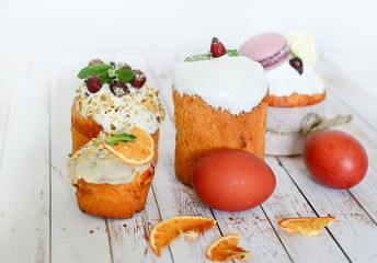 Easter cakes and multi-colored eggs decorated with macaroons, mint, wild rose with white icing, lilac branches on a white wooden background. Easter.