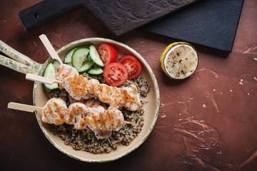Fried chicken fillet on skewers, quinoa and fresh vegetables in a serving pan, top view over rusty metal background