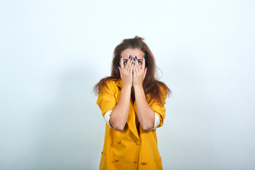 Attractive young woman in yellow jacket covering eyes, looking through hands isolated on gray...