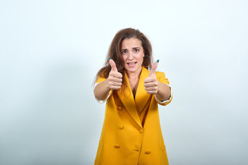 Young caucasian attractive woman in yellow jacket keeping thumbs directly isolated on gray...