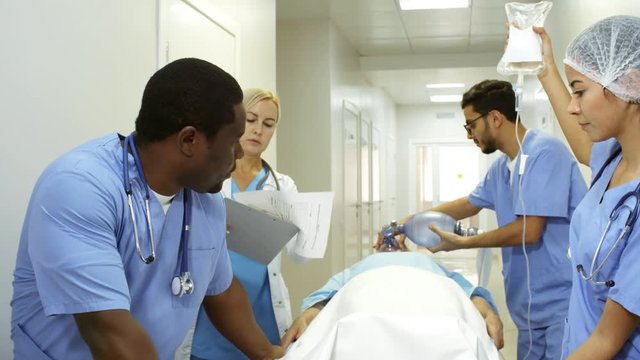 Dolly shot of multi-ethnic group of emergency room nurses in scrubs pushing gurney and administering oxygen and IV fluids to unresponsive patient as female doctor with clipboard giving instructions