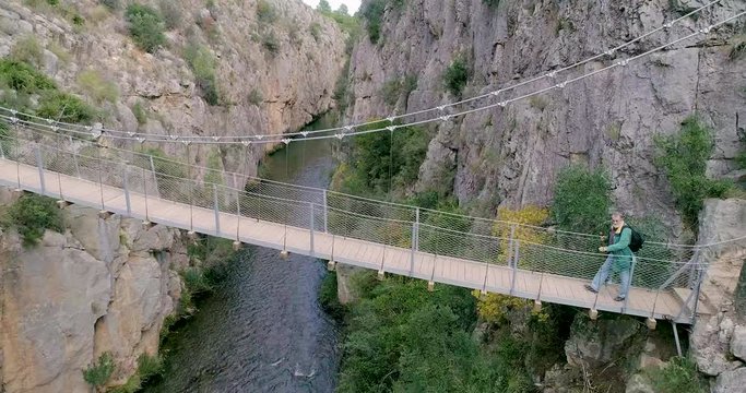 Dron en movimiento lateral de puente colgante y excursionista senior cruzando