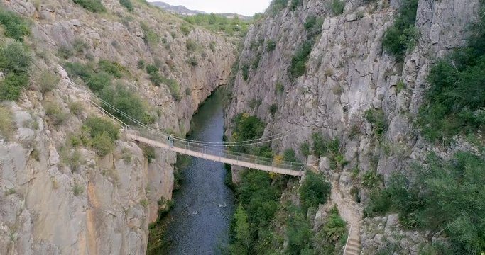 Senderista anciano cruzando un puente colgante sobre el abismo y el dron acerc&aacute;ndose lentamente