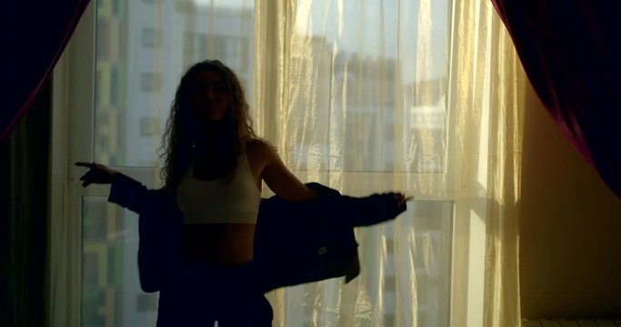 a curly-haired young beautiful girl who is standing in front of a large window, wearing a white short top, black pants and a jacket, she poses and touches her hair, makes dancing movements.