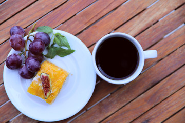 Blurred food background. On a wooden table is a white plate with a piece of yellow cake with a bunch of blue grapes and a cup of coffee. Close-up, top view, horizontal. Food and holiday concept.