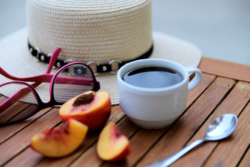 Blurred food background. On a wooden table is a white cup with coffee and peach slices. On the side plan is a hat and sun glasses. Close-up, side view, horizontal. Food concept.